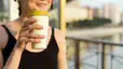 A close-up photo of a woman drinking coffee after a workout. She's standing outside and holding a reusable to-go cup.