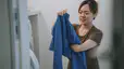 A woman holding up and folding a blue long-sleeve shirt at home in the laundry room.