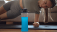 A blue sports drink in the foreground with a woman doing a side plank on a workout mat in the background.