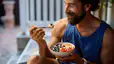 A man eating a bowl of oats topped with fresh fruit while sitting outside. He's eating carbs before his workout.
