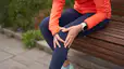 A woman experiencing runner's knee. It's a close-up photo of her sitting on a park bench after a run and holding onto the front of her left knee.