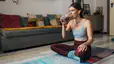 A millennial woman sitting on a yoga mat at home and drinking a glass of chocolate milk after a workout.