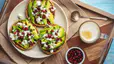 A plate of flat circular pieces of bread with avocado, pomegranate seeds, and feta cheese with a latte and extra bowl of pomegranate arils on the side.