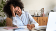 A mentally exhausted woman sitting at her desk in front of her laptop. She's holding her head in one hand and her glasses in another. She looks stressed out and tired.