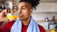 An athlete drinking a glass of orange juice before going on a run. He's in his kitchen and has a towel around his neck.