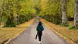 A woman walking down a road surrounded by trees in autumn. Learn the mental health benefits of walking in this article.