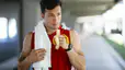 An athlete eating a banana after a workout while walking outside. He is wearing a red athletic tank top and has a white sweat towel over his shoulder.