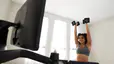 woman doing a shoulder press with dumbbells next to a Peloton Tread