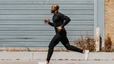 A man on a long run outside. He's wearing all black and jogging in front of an industrial building.