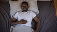 A man practicing breathing exercises for sleep. He's lying on his back in bed, eyes closed,  with his right hand on his upper abdomen.