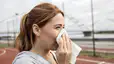 In this image for an article about whether or not you should exercise when you're sick, a woman is blowing her nose into a tissue while she stands outside on a sports court of some kind.