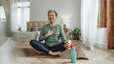A woman meditating on an indoor yoga mat, with one hand on her chest and the other on her knee, to practice self-care during menopause.