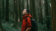 A man hiking in a dense forest, looking up at the sky.