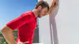 A tired male athlete leaning against a white wall outside. His face is red, with one hand on his hip and another arm leaning against the wall. He's resting his head against his arm and wearing a red T-shirt. It's a blue, sunny day outside.