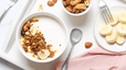 An array of high-protein snacks on a white table, such as Greek yogurt, bananas, and nuts.