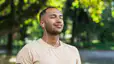 A man practicing mindfulness and meditation. He is closing his eyes and smiling while listening to wired headphones in an outdoor setting.
