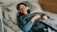 A relaxed woman meditating in bed. She has headphones on and is under a blanket.