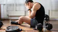 A man dealing with exercise burnout. His head is in his hands and he's feeling mentally drained, surrounded by dumbbells and weights.