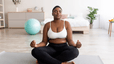 A woman sitting on a yoga mat at home doing a postpartum workout. She is in lotus position with her eyes closed.