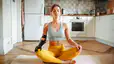 A woman meditating on her yoga mat at home in her kitchen. She's sitting in lotus position, closing her eyes, and has her hands resting on her knees.