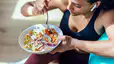 A woman eating dinner (a veggie-packed grain bowl) before or after a workout. She's in workout clothes and is leaning against a big exercise ball. 
