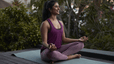 A woman meditating in a seated meditation position. Her hands on resting on her knees, she's sitting up tall, and she's smiling with her eyes open.