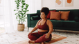 A woman practicing healthy habits by meditating at home. She's wearing activewear and sitting on a blanket on the floor in a sunny living room.