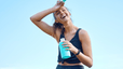 A happy athlete rubbing sweat from her forehead and drinking from a teal water bottle while standing outside. Learn about the benefits of electrolytes here.