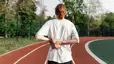 A photo of the back of an athlete who is on an outdoor track. They are feeling bloated after a workout and holding their back with their hands. 