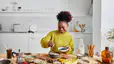 A happy woman at home preparing a healthy breakfast with complex carbohydrates like fruits and nuts.