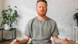 A man practicing a meditation for stress at home in a bright, sunny room. His eyes are closed and his hands are resting on his knees as he sits in a cross-legged position.
