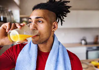 An athlete drinking a glass of orange juice before going on a run. He's in his kitchen and has a towel around his neck.