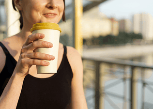 A close-up photo of a woman drinking coffee after a workout. She's standing outside and holding a reusable to-go cup.