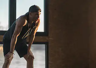 A tired athlete with his hands on his knees, bent over. He's experiencing shaking after a workout.