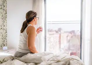 A woman sitting in bed drinking water first thing in the morning as she looks out the window.