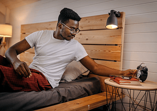 A man going to bed, looking at his alarm clock while sitting under the covers.