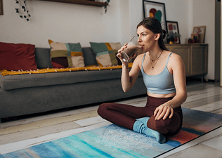 A millennial woman sitting on a yoga mat at home and drinking a glass of chocolate milk after a workout.