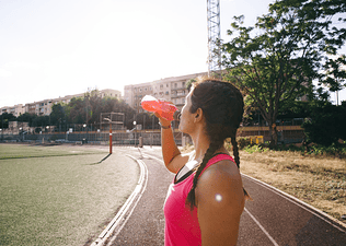 A side view of a woman drinking an orange-red electrolyte drink on an outdoor track.