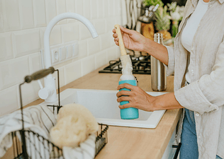 A woman cleaning a water bottle using a brush in the kitchen sink.