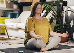 A woman meditating in lotus position, sitting with her eyes closed on a yoga mat at home. Learn about the benefits of meditating before or after a workout in this article.