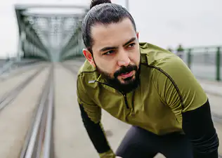 A man exhaling as he puts his hands on his knees while standing up outside. He's dealing with a stomach ache after exercise.