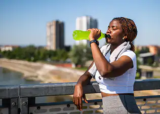 A runner leaning against a fence and drinking a green sports drink to replenish electrolytes on during a hot, sunny run.