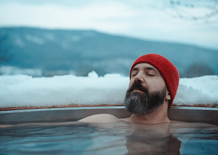 A man resting in a hot tub in a snowy outdoor environment. He's wearing a red beanie.