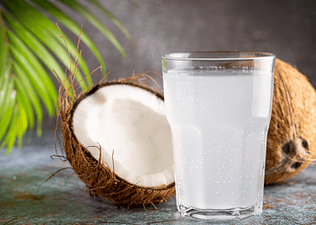 A clear glass of coconut water sitting in front of coconuts. There are many health benefits of coconut water.