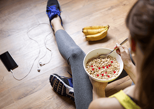 A woman eating oatmeal and a banana before going on a morning run. Her phone and headphones are on the floor next to her.