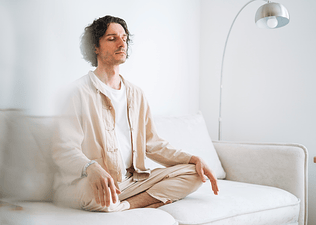 A young man sitting cross-legged on a white couch in a bright room and meditating.