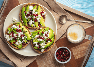 A plate of flat circular pieces of bread with avocado, pomegranate seeds, and feta cheese with a latte and extra bowl of pomegranate arils on the side.