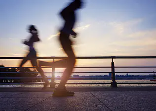 The silhouettes of two runners on a waterside path during sunrise to illustrate the concept of running twice a day.