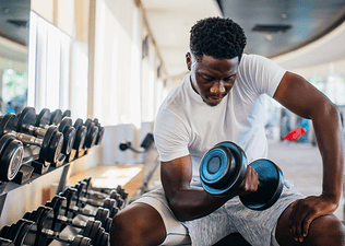 A man sitting at the gym curling a dumbbell with one arm while sitting on a workout bench.