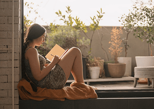 A woman writing affirmations for self-esteem in her journal. She's sitting on her apartment terrace on a nice day, leaning against the open doorway.